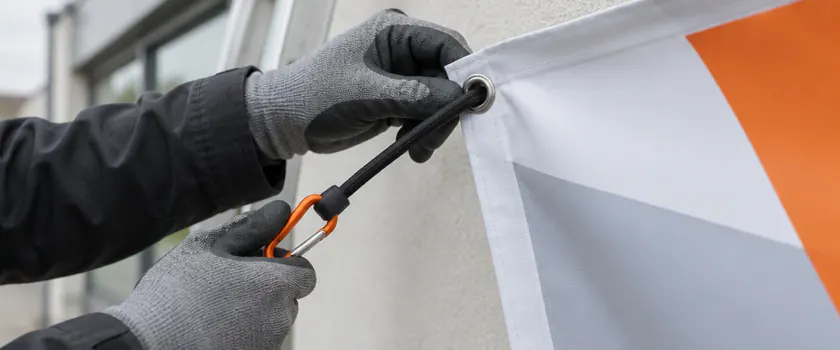 Dłonie w rękawicach przewlekają linkę przez oczko w rąbku banera napiętego na elewacji budynku.